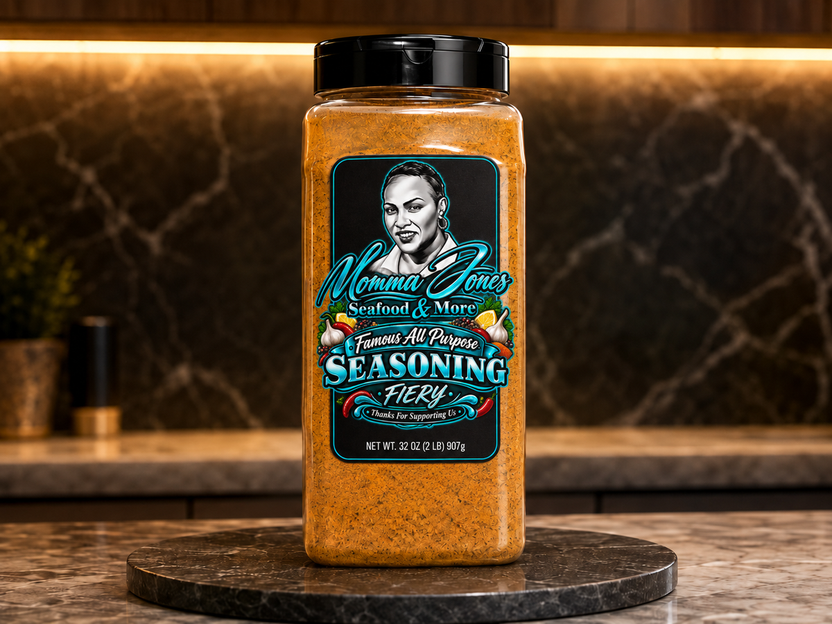 A large plastic jar of Momma Jones Seasoning Fiery stands on a round dark stone coaster in a kitchen setting. The jar is filled with a bright orange-brown seasoning blend and has a black lid with a prominent black, teal, and white label featuring a portrait of Mamie Jones.